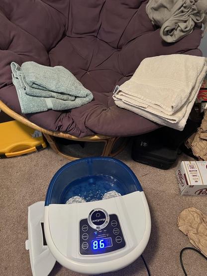 A blue and while foot bath bubbling away as it heats up, in front of a purple corduroy mama San chair with two large, fluffy bath towels folded for later use.