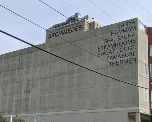 Fachada de un edificio de concreto, sin ventanas, con cables colgados en frente, que lleva el texto: Archimedes...