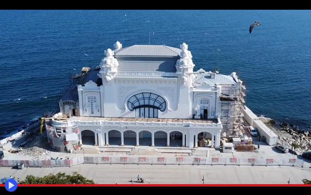 Foto architettonica di edificio in stile liberty sul lungomare, con un portico elegante e grande vetrata a forma di conchiglia. Ai margini del quadro prospettico, è possibile intravedere il materiale accatastato di un antistante cantiere.