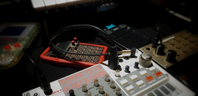 Photo with Blur and Vignette.

Some connected gear on a black cloth.
In the background a clear Game Boy and a Teenage Engineering PO-28 Robot with an old red iPhone case. Left to it a Korg Mini Kaoss Pad 2S.
In the foreground on the right a Volca Keys and in the front a Volca Sample. It has a grey sticker with red text in the top middle, almost matching the grey/white case:

Playing Yo-Yo Is Not A Crime