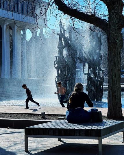 Two boys in silhouette run across a public fountain while a woman seated on a bench minds her own business.