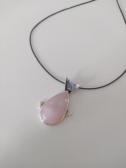 a fish shaped silver pendant with a teardop shaped pink gemstone as the main body of the fish. it has a black cord