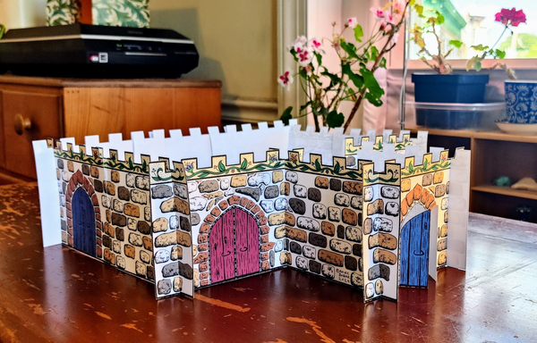 A photo of a 3D paper castle made of sections of white paper printed with stones, crenellation with a flowering vine motif, and doors, all in black ink. The castle is viewed at an angle and from slightly above. Some of the stones are colored in in shades of light yellow-beige and dark grey-brown, and the crenellation is a warm yellow. The vine pattern in the crenellation has been colored green, and the flowers are colored in a variety of shades of blue, purple, pink, and orange. The castle shape is a large rectangle, with a smaller square room extending off the front. At the front of the smaller room is a set of large blue double "wooden" doors set inside a reddish-brown stone gothic arch. On the front wall of the main castle to the left of the smaller room is another set of large double "wooden" doors, this time colored red. Around the corner at the rear of the left side wall of the castle is a third set of double "wooden" doors, colored blue again. The castle sits on a scratched wooden desk. Behind and to the left, a black scanner sits on a wooden bureau. A potted geranium with pink and white flowers is visible behind the castle. Behind and to the right of the castle is a window with another smaller geranium, and an assortment of tchotchkes in a bamboo box on the windowsill.