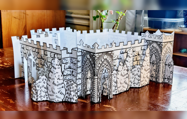 A photo of a 3D paper castle made of sections of white paper printed with stones, crenellation, and doors in black ink, viewed from above and at an angle. The castle is a simple square shape, with a smaller square room extending off the back of the castle. There are two slightly different porticos on either side of the front wall, with crenellated tops and shrubbery between. The portico on the left has pointed gothic arches on all three sides, but no door except on the interior wall. The portico on the right has doors on all three sides, set into the same gothic arches; the one in front is folded so it stands ajar. Each stone arch contains decorative vines from top to bottom, with a tiny moon at the pinnacle: waxing crescents on the left side arches, full moons in front, and waning crescents on the right. The wooden doors inside each arch of vinework on the right side portico are shaped like a pointed gothic arch with elaborate iron hinges. The stone arches of both porticos are framed by an arch of gravelly stucco, but this area is very light on the right side portico, and much darker on the left side portico. Above this on both porticos, many small round river stones are set into the stucco, with a small inverted triangle of stone bricks at each corner. More shrubbery wraps around the left side of the castle towards a set of large plain wooden doors standing ajar inside a stone gothic arch. The castle sits on a scratched wooden desk.]