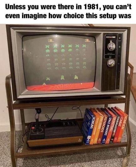 Captioned photograph of a decent-sized old color CRT television with analog dials, showing a game of Space Invaders being played on an Atari home gaming console. There are at least 10 different game cartridges stacked beside the console. Caption reads "Unless you were there in 1981, you can’t even imagine how choice this setup was."
