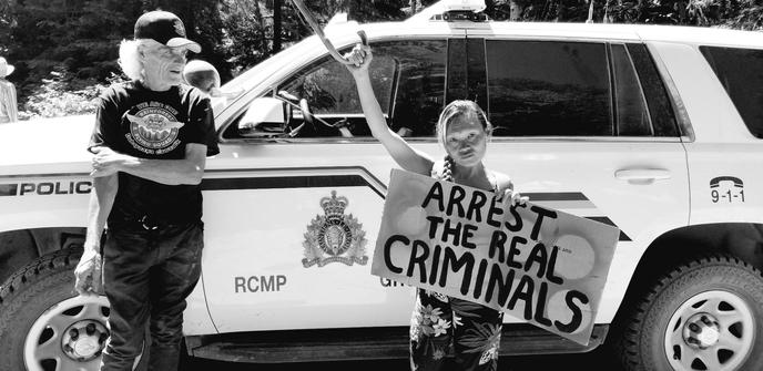 Me, standing in front of an RCMP SUV, with my right hand holding my wooden cane up & holding a sign in other hand that says: ARREST THE REAL CRIMINALS

Black and white photo.
