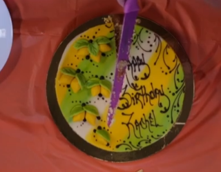 A beautifully decorated cake with marzipan lemons being cut with a bright purple knife. Happy Birthday Rachel is written on top.
