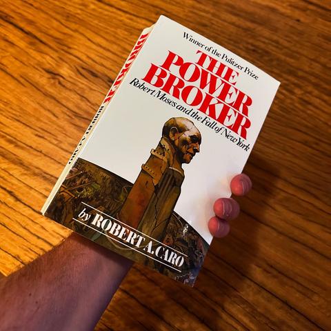 A hand holds the book "The Power Broker" by Robert A. Caro, featuring a cover illustration depicting a bust of Robert Moses. The book is set against a wooden background.