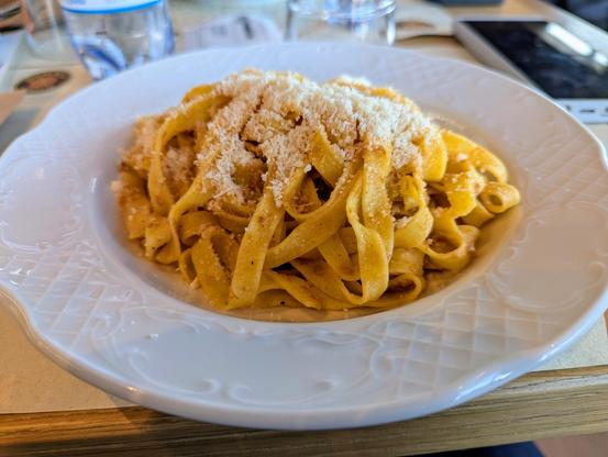 A plate of tagliatelle pasta topped with grated cheese, served on a white ceramic dish. The pasta appears to be lightly seasoned with a golden sauce. The background includes a water bottle, some paper, and a smartphone placed on the table.