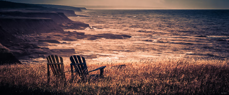 A pair of adorondak style chairs in a grassy field overlooking the ocean from a cliff in evening light.