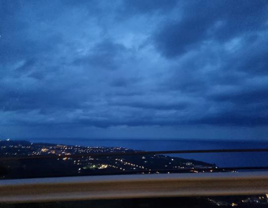 Città vista dall'alto con le luci accese di sera, mare blu intenso e cielo blu carico di pioggia