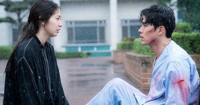 Park Shin Hye in casual clothes & Hyun Bin in hospital clothing with blood on his arm & face, sitting on the ground in the rain looking at each other. From the 2018 South Korean drama "Memories of the Alhambra."