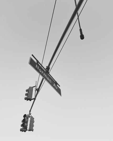 View from below of a traffic sign post from which several stoplights and a sign reading “Norman Av” hang, in black and white.