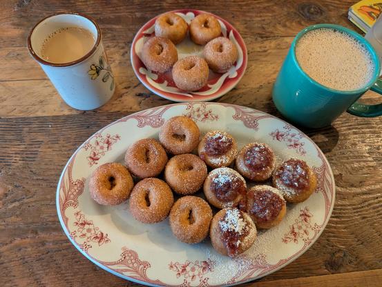 Two plates of small, silver dollar-sized doughnuts and chai. The bottom plate has six cinnamon sugar doughnuts, and six maple bacon. The top plate has just five cinnamon sugar doughnuts. Next to the plates on the wooden table is a cup of chai and a cup of coffee.
