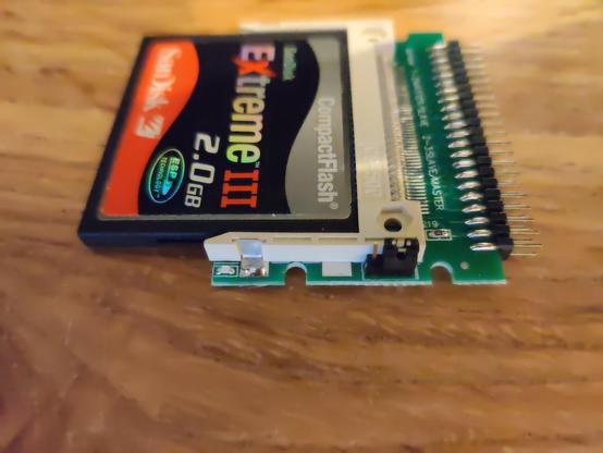 A side-on macro photo of a Compact Flash to 40-pin IDE adapter sitting on a wooden table, with a SanDisk Extreme III 2.0GB card inserted. Along the edge is a jumper across the two rightmost of three pins. The CF/IDE card works in this mode.