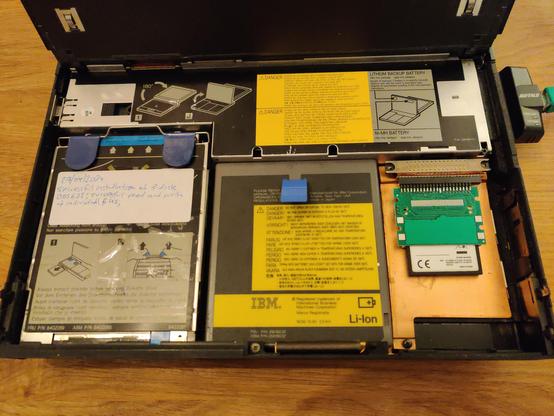 A 45° downward view into the Thinkpad 760CD with the bonnet up (actually the keyboard) showing the superbly modular design. At the front left is a floppy disk drive (complete with sticker outlining how I've exercised it recently), in the middle the still-working battery, and on the right, the hard drive bay with the CF/IDE adapter connected to the proprietary internal connector and the underside of a Compact Flash card visible. (The adapter gets installed face-down.)