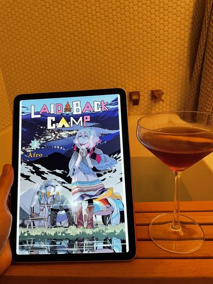 An iPad showing the cover to volume 2 of the manga “Laid-Back Camp”, a Manhattan cocktail, both on a small wooden platform over a bathtub