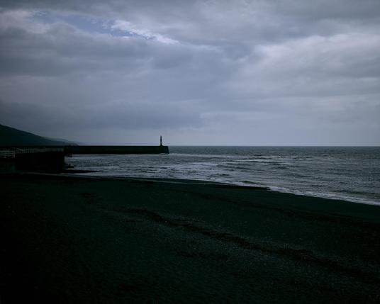 The image depicts a coastal scene on a cloudy day. The foreground shows a dark beach, while gentle waves break on the shore. A long, straight pier extends into the sea from the left, with a faint silhouette of a lighthouse at the end. The sky is overcast with dense, gray clouds.