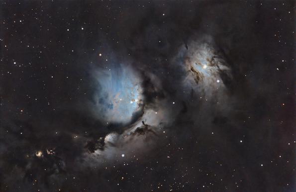 Blue reflection nebula with two main areas one in the center of the frame, with a dark cloud cutting through the middle. the other is to the right, and has several dark wispy clouds in front of it. The nebula is survived by a field of stars.