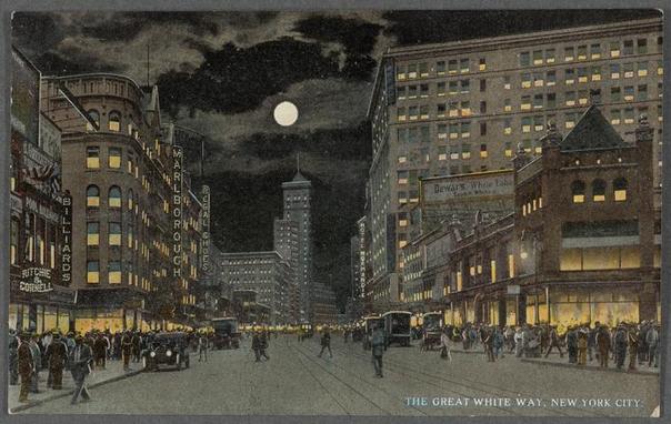 The image is a vintage postcard depicting a bustling city street at night. The scene appears to be from the early 20th century, based on the style of buildings and vehicles. Large signs with illuminated text adorn various commercial establishments lining both sides of the wide avenue. Notable among them are "Marlboro," "Rodgers & Hammerstein," "Bull's Head Inn," and "Dewey's White Label." The architecture is indicative of early skyscraper design, with numerous tall buildings featuring bright window illuminations suggesting active businesses within.

The street below is filled with pedestrians in period attire, indicating a lively urban environment. In the midst of this scene are several horse-drawn carriages, reflecting pre-motorized transport methods typical for that era. A trolley or tram can be seen further down the avenue, adding to the sense of movement and activity.

The sky above is dark with clouds partially obscuring what seems to be a full moon, casting an eerie glow over the cityscape below. The overall mood conveyed by this image suggests a vibrant yet somewhat mysterious urban nightlife from a past era.