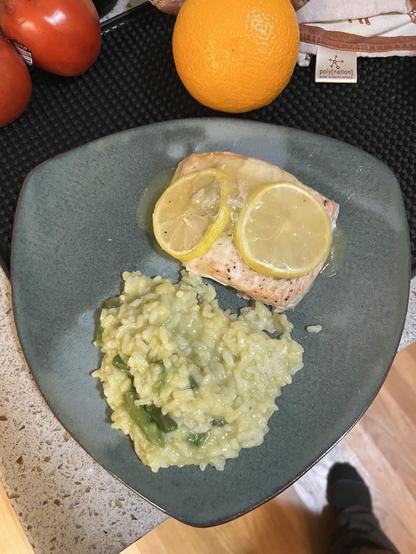 Salmon filet topped with lemon slices and lemon-butter sauce, plated alongside asparagus risotto on a triangular seafoam green plate. There is an orange and a tomato in the background on the cabinet.