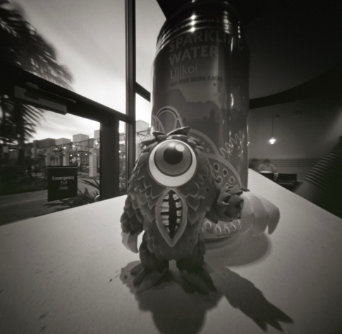A one eyed monster figurine on a table in front of a drink can, taken with a pinhole camera