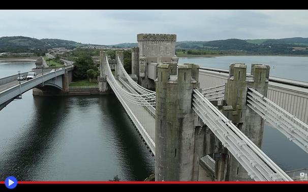 Veduta prospettica con tre ponti che attraversano parallelamente un ampio fiume: stradale, pedonale e ferroviario. Due su tre presentano torri con le riconoscibili caratteristiche di un piccolo castello medievale.