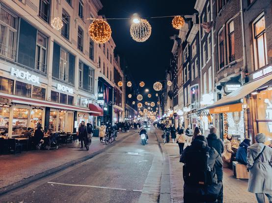 Street in Amsterdam bustling with pedestrians and cyclists at night, adorned with festive spherical lights hanging above. Neon signage for "Hotel Doria" and "Dam Apotheek" is visible. People dine outdoors and walk along the brightly lit shops, creating a lively urban ambiance.