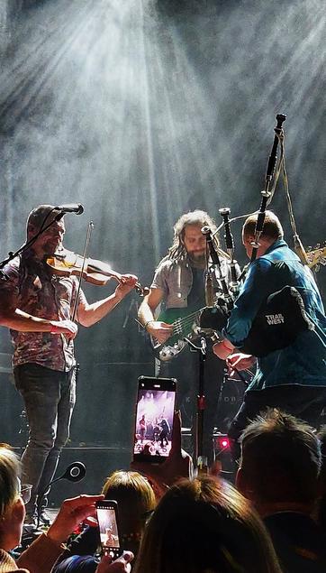 Photo of three musicians from the band Skerryvore performing on stage. Left the fiddle, in the middle the Bass and right the bagpipe. The three musicians facing each other.