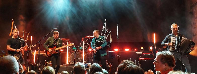 Photo of four musicians from the band Skerryvore performing on stage. From left to right: bagpipes, guitar, bagpipes and accordion. The musicians dancing on stage.