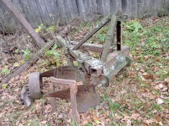 A rusty old plow with two blades. A spare wheel lays next to it. The actual, attached wheel is hidden by the frame. Some other steel parts are leaning against it. Some lichen is growing on it, too.