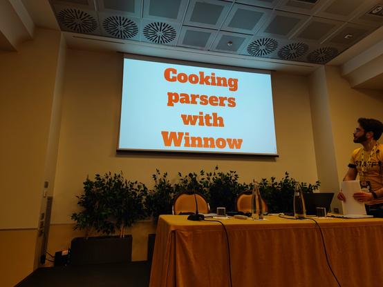A screen showing the title slide "Cooking parsers with Winnie", in front of a yellow wall. Before the screen is a desk with two chairs and one of the organizers of RustLab in a yellow shirt.