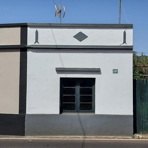 Photograph of a single-storey house facade in the Canary Islands featuring a horizontal rhombus shape on the middle and two vertical bifurcated arrows on the sides of the frieze section of the facade. The house has a single window overlooking the street, the sky in the background is bright blue.
