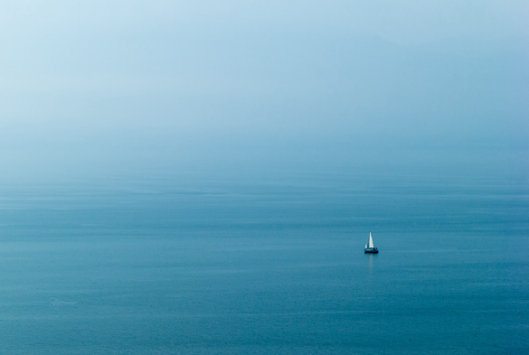 Das Foto zeigt eine ruhige, minimalistische Szene auf offenem Wasser, das in verschiedenen Blautönen schimmert. Die Weite des Wassers ist fast leer, bis auf ein einzelnes Segelboot, das sich im unteren rechten Bereich des Bildes befindet. Der Himmel und das Wasser verschmelzen im Hintergrund durch die sanften Blautöne und das sanfte Licht. Die Atmosphäre wirkt friedlich und leicht melancholisch.