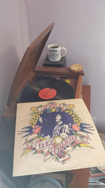 Record player with a 12" black vinyl record. In the foreground is an album cover with a tattoo like pattern with Rory Gallagher in the centre. Behind the player is a mug of coffee on a speaker.