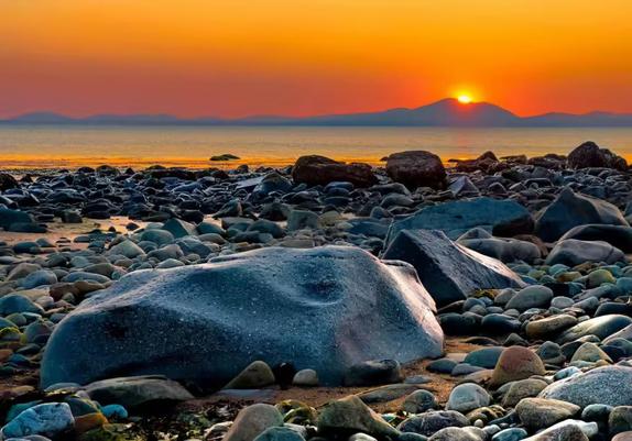 Large stones on a beach in the foreground; in the background the sun sets behind a range of hills. The sea lies between foreground and background.