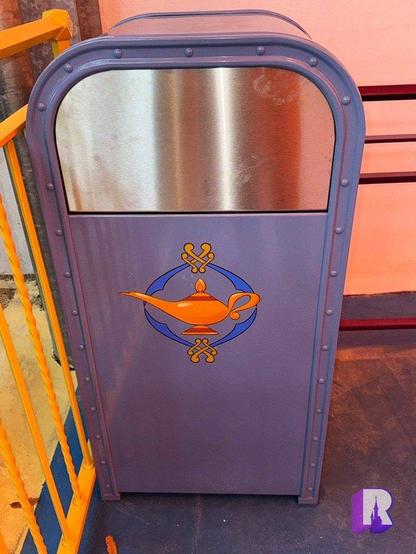 A decorative trash can with a light purple body located in a theme park. It features a gold lamp design, reminiscent of an oil lamp, surrounded by a circular emblem.
