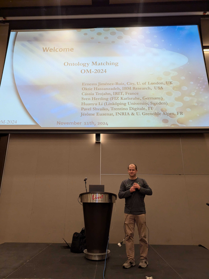 Sven Herling standing in front of the Big Screen at the Ontology Matching Workshop 2024 at ISWC 2024 in Baltimore, USA. The Screen is showing Welcome to Ontology Matching OM-2024 and lists all the responsible co-organisers: 

    Ernesto Jiménez-Ruiz
    City, University of London, UK & SIRIUS, University of Oslo, Norway
    Oktie Hassanzadeh
    IBM Research, USA
    Cássia Trojahn
    IRIT, France
    Sven Hertling (Main contact)
    FIZ Karlsruhe, Germany
    Huanyu Li (Main contact)
    Linköping University, Sweden
    Pavel Shvaiko
    Trentino Digitale S.p.A., Italy
    Jérôme Euzenat
    INRIA & Univ. Grenoble Alpes, France