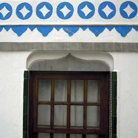 Una foto de una fachada de una casa canaria en Fuerteventura con características cenefas azules similares a montañas en un reborde por encima de la ventana y otro reborde superior con símbolos azules en forma de estrella de cuatro puntas dentro de un círculo, repetidos en una lína horizontal.