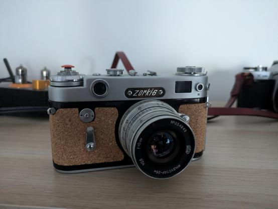A picture of the Zorki-6 rangefinder with an orange shutter, a cork body, and a relatively large 50mm lens.  In the background, an analog vacuum tube audio interface and a Pentax K1000 are visible.