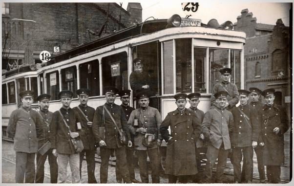 Zu sehen ist ein altes Schwarz-Weiß Foto, wahrscheinlich aus den 20er oder 30er Jahren. Auf dem Bild sind hintereinander zwei Wagen einer alten Straßenbahn zu sehen. Der vordere Wagen hat die Nummer 29 und ein Schild auf dem Hannover Bahnhof steht. Vor dem Wagen stehen insgesamt 15 Männer in der Uniform und mit der Mütze eines Straßenbahn-Schaffners oder -Fahrers. Einige der Männer haben Kassierbeutel umgehängt. Im Bildhintergrund ist ein gemauerter Lockschuppen erkennbar.
