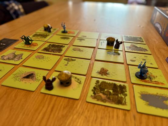 A tabletop game board comprised of 25 tiles that feature images of grass, trees, flowers, mountains, and a lake. On the board are miniatures of cottages, peasants, knights, and an archer.