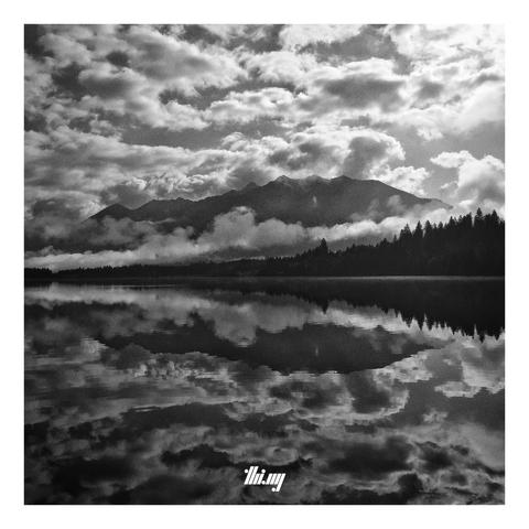 Grainy B&W photograph of a completely still lake reflecting the large Karwendel mountain massif, the silhouette of the forest in front of it and the dozens of small clouds surrounding it all (incl. a band of fog floating just above the forest). The reflections on the water surface are much darker. A sliver of sunlight hits the water just near the edge of the forest...