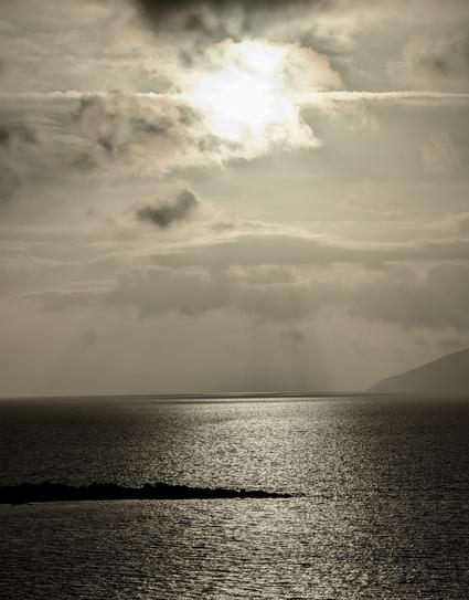 A photo looking over the sea towards the Isle of Rum, Scotland with the sun high in the sky and very bright but hazy clouds make it look dreamy.