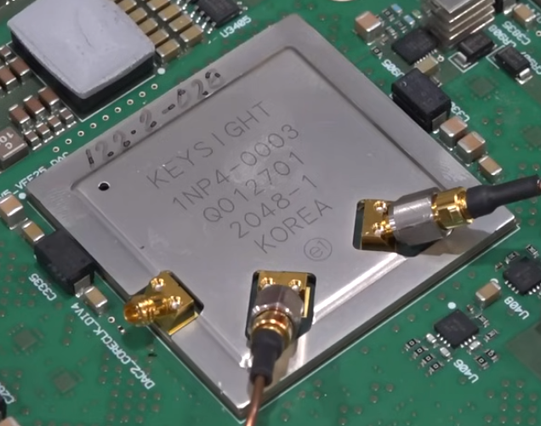 A large-ish BGA package on a green circuit board surrounded by various parts, mostly surface mount ceramic capacitors. The lid has cutouts for three angled coaxial RF connectors.

On top side of the package, 122-2-028 is written using black marker.
The package itself is labeled
KEYSIGHT
1NP4-0003
Q012701
2048-1
KOREA