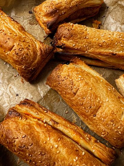 A close-up of several freshly baked vegan chorizo sausage rolls sprinkled with sesame seeds on a sheet of parchment paper. The pastries have a golden-brown color and a flaky texture.