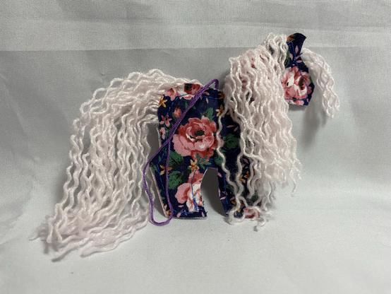 A crafted horse figure made from fabric featuring floral patterns, with curly white yarn representing its mane and tail. The horse stands on a light background.