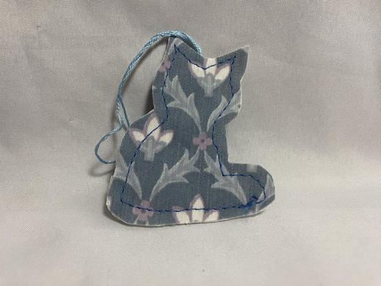 A fabric cat silhouette in shades of gray with floral patterns, stitched with blue thread and featuring a small hanging loop.