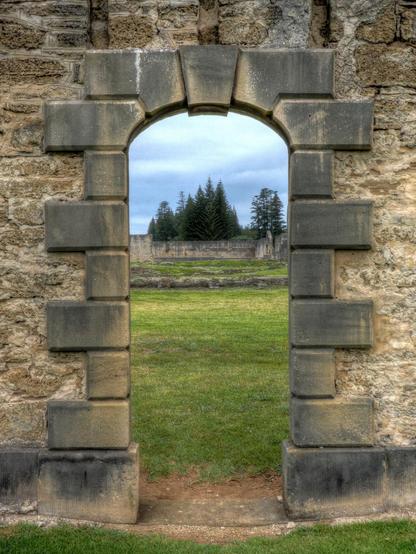 Looking through one of the stone-edged portals into the remains of the prison compound. Thick green grass covers the ground, the remains of wall foundations peek through, and at the far end, the exterior wall remains, with pine trees and the sky beyond.