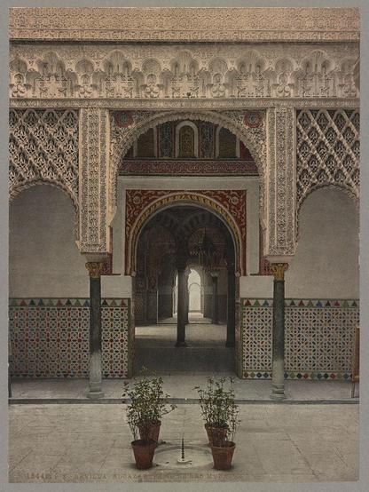 The image depicts an architectural interior scene of a historic building. It shows the entrance to what appears to be a grand hall or courtyard, featuring intricate and ornate decorations typical of Islamic artistry found in Spanish Moorish architecture.

In the foreground, there are two potted plants on either side of what looks like an archway leading into a deeper space within the structure. The walls surrounding these planters have colorful geometric patterns reminiscent of traditional Andalusian designs with vibrant hues such as reds, blues, greens, and yellows.

Above is a series of white arched windows or passageways that allow light to filter through, casting soft shadows on the floor below. These archways are part of a larger decorative scheme incorporating elaborate floral motifs in alternating bands of color along their edges.

The center focal point is an ornately carved wooden door with red and gold embellishments indicating significant craftsmanship and cultural importance within this context. It's framed by two white columns which support a canopy or portico above the doorway, adding to its majestic appearance.

This particular photograph captures a moment in time at Seville Alcazar around 1890-1906 as per an inscription visible on one of the lower corners where it says "Sevilla Alcazar," suggesting that this was taken within or near what is now known as Royal Palace of Seville. The overall ambiance evokes [...]
